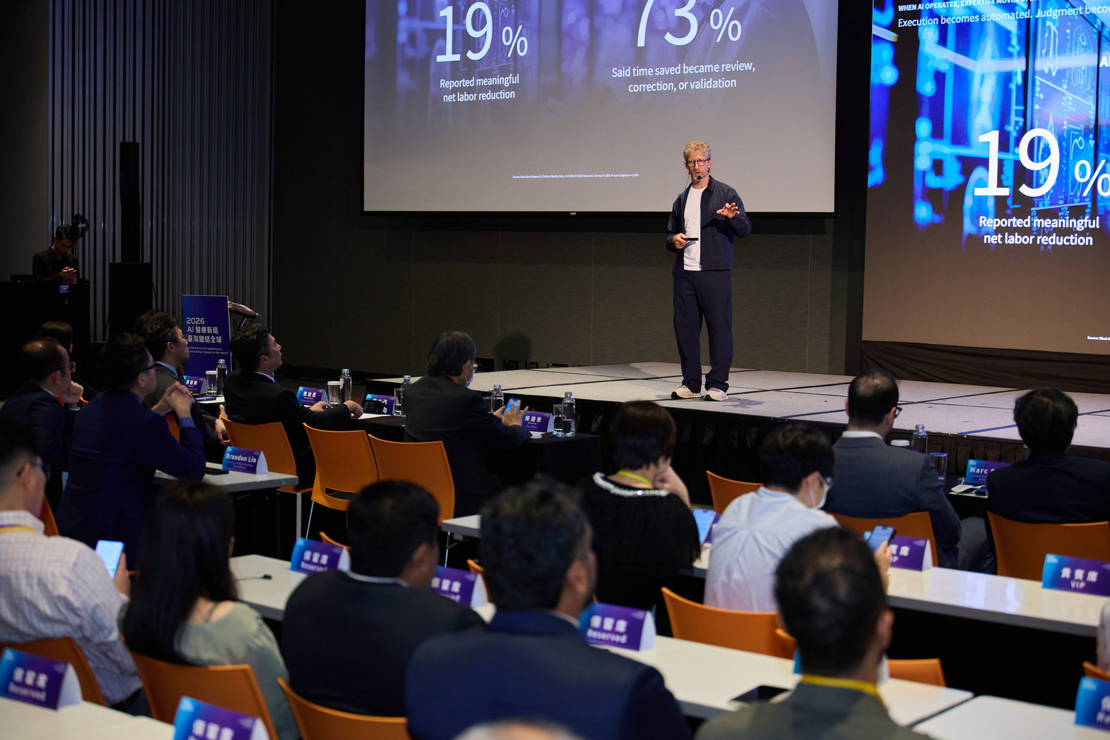 Jan Beger keynoting at A New Era of AI Healthcare in Taiwan, beside a slide reading 19% reported meaningful net labor reduction